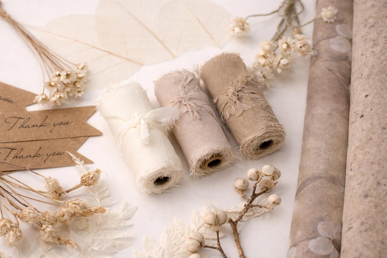 May include: Three rolls of frayed fabric ribbon in cream, blush, and tan, with dried floral accents and "Thank you" tags. The ribbons are tied with matching fabric. Rolls of patterned paper are in the background.