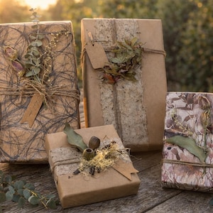 May include: Four wrapped gifts with natural brown paper and twine. The gifts are decorated with dried leaves, sprigs, and tags. The gifts are arranged on a wooden surface with greenery in the foreground, with a blurred background.