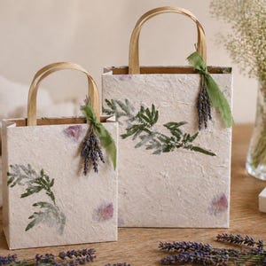 May include: Two rectangular gift bags made of textured off-white paper, each featuring green botanical leaf imprints. The bags have light brown paper handles and are decorated with a small bundle of dried purple lavender tied with a green ribbon.