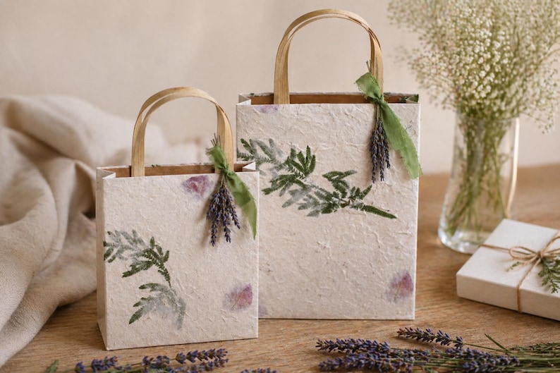 May include: Two rectangular gift bags made of textured off-white paper, each featuring green botanical leaf imprints. The bags have light brown paper handles and are decorated with a small bundle of dried purple lavender tied with a green ribbon.
