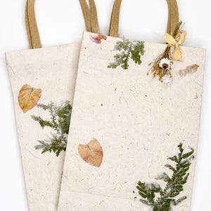 May include: Two rectangular gift bags made of off-white textured paper with brown paper handles. The bags are decorated with pressed green fern leaves, dried orange leaves, and a small bundle of dried white flowers tied with a light beige ribbon.