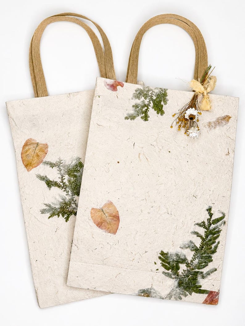 May include: Two rectangular gift bags made of off-white textured paper with brown paper handles. The bags are decorated with pressed green fern leaves, dried orange leaves, and a small bundle of dried white flowers tied with a light beige ribbon.