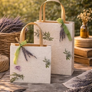 May include: Two rectangular off-white paper gift bags with flat handles, decorated with pressed green foliage and purple flower petals. Each bag is tied with a green ribbon and a small bundle of dried lavender stems attached to the handle.