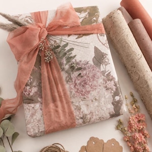 May include: A rectangular gift box wrapped in floral patterned paper featuring muted pink and brown botanical illustrations. The package is tied with a wide, sheer terracotta-colored ribbon and decorated with dried pampas grass and eucalyptus sprigs.