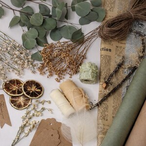 May include: An assortment of craft supplies including dried flowers, eucalyptus leaves, twine, paper rolls, and tags. The color palette is neutral with shades of brown, green, and cream. Some tags read "Thank you".