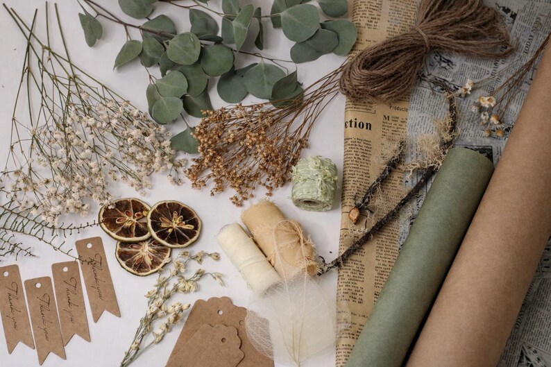 May include: An assortment of craft supplies including dried flowers, eucalyptus leaves, twine, paper rolls, and tags. The color palette is neutral with shades of brown, green, and cream. Some tags read "Thank you".
