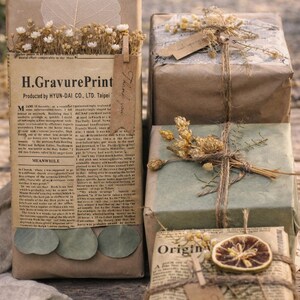 May include: Several wrapped gifts with brown paper, twine, and dried floral accents. One package features newspaper print and the text "H.GravurePrint". Another has a tag that says "Thank you". The gifts are arranged on a wooden surface.