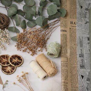 May include: An overhead shot of craft supplies including dried orange slices, twine, dried flowers, eucalyptus leaves, rolls of paper, and rolls of ribbon. There are also brown tags with the words "Thank you" printed on them.