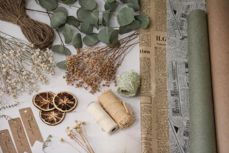 May include: An overhead shot of craft supplies including dried orange slices, twine, dried flowers, eucalyptus leaves, rolls of paper, and rolls of ribbon. There are also brown tags with the words "Thank you" printed on them.