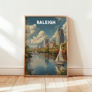 May include: A framed art print featuring a scenic view of Raleigh, North Carolina. The artwork depicts a sailboat on a river, with city buildings and colorful trees in the background. The word "RALEIGH" is displayed at the top in white letters.