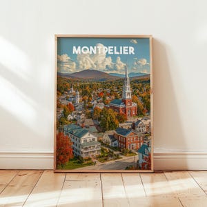 Op de afbeelding: Een ingelijste print met een luchtfoto van Montpelier, Vermont. De afbeelding toont een stad met een kerk en een torenspits, omgeven door herfstbladeren. De lucht is blauw met witte wolken. Het woord "Montpelier" staat bovenaan afgedrukt.