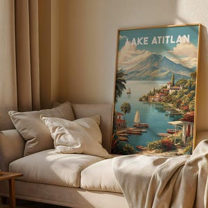 May include: A framed art print titled "Lake Atitlan" shows a scenic landscape with a lake, mountains, boats, and buildings. The artwork has a gold frame and is displayed on a beige sofa with pillows and a blanket.