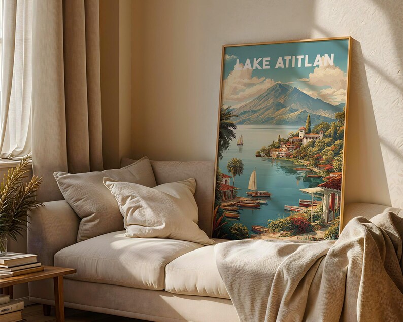 May include: A framed art print titled "Lake Atitlan" shows a scenic landscape with a lake, mountains, boats, and buildings. The artwork has a gold frame and is displayed on a beige sofa with pillows and a blanket.