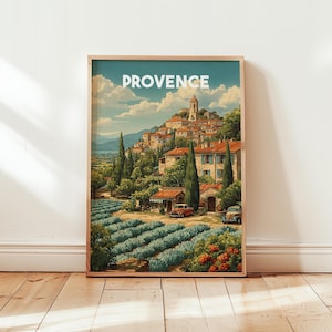 Op de afbeelding: Een ingelijste kunstafdruk van een schilderachtig dorp in de Provence, Frankrijk. De prent toont een heuvelstad met rode pannendaken, weelderig groen en het woord "PROVENCE" bovenaan. Een oldtimer staat geparkeerd voor een gebouw.