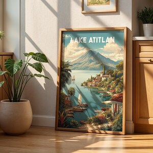 May include: A framed art print depicting Lake Atitlan, showcasing a colourful landscape with a mountain, lake, and village. The print is in a wooden frame and leans against a wall. The scene is illuminated by sunlight, with sailboats on the water.