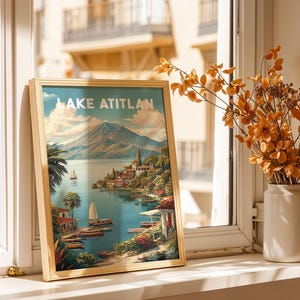 May include: A framed print of Lake Atitlan, depicting a scenic view of the lake, mountains, and buildings. The print is in a light wooden frame and is propped up on a windowsill. A vase of orange flowers sits nearby.