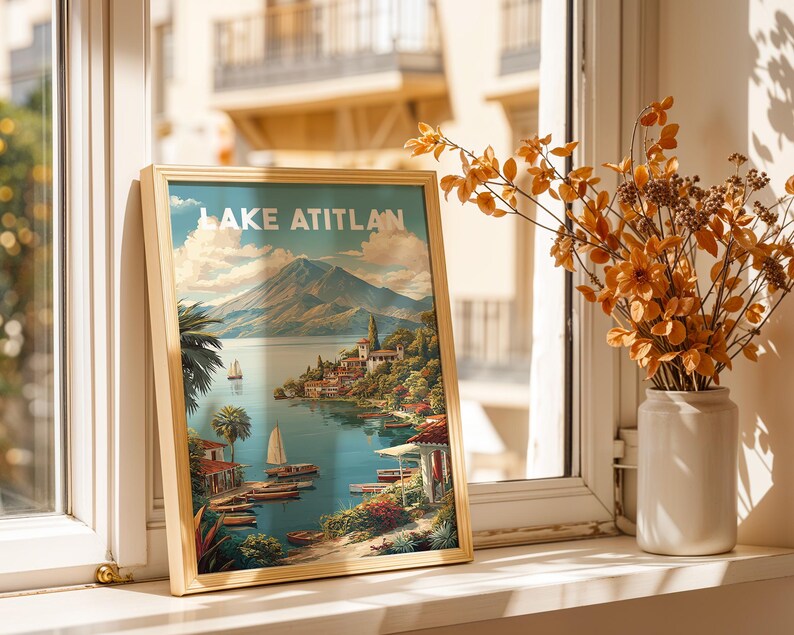 May include: A framed print of Lake Atitlan, depicting a scenic view of the lake, mountains, and buildings. The print is in a light wooden frame and is propped up on a windowsill. A vase of orange flowers sits nearby.