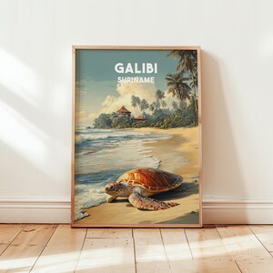 Op de afbeelding: Een ingelijste kunstdruk met een strandscène met een zeeschildpad op het zand. De print heeft de tekst "GALIBI SURINAME" bovenaan. Het kunstwerk toont een tropisch strand met palmbomen, oceaangolven en een gebouw op de achtergrond.