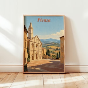 Op de afbeelding: Een ingelijste kunstdruk met een schilderij van het stadsplein van Pienza, Italië. De afbeelding toont een stenen kerk met een hoge klokkentoren, gebouwen in terracottakleur en een uitzicht op glooiende heuvels onder een helderblauwe lucht.