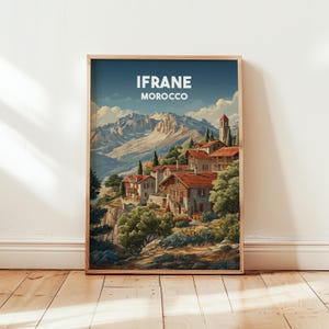 Könnte beinhalten: Ein gerahmter Druck mit einer malerischen Ansicht von Ifrane, Marokko. Das Kunstwerk zeigt ein Dorf mit roten Ziegeldächern inmitten üppigen Grüns und Bergen unter blauem Himmel. Der Text "IFRANE MAROKKO" ist oben zu sehen.