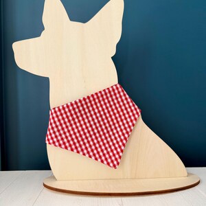 May include: A wooden dog-shaped decoration with a red and white checkered bandana. The dog silhouette is light-colored wood, mounted on a round wooden base. The bandana adds a pop of color to the neutral tones.