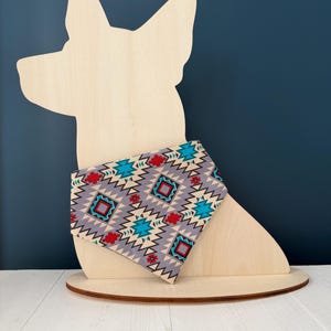 May include: A wooden dog-shaped display with a patterned bandana. The bandana features a gray background with red, turquoise, and white geometric designs. The display is on a white surface against a dark blue backdrop.