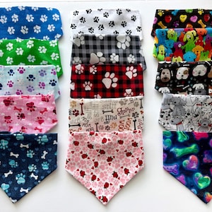 May include: A collection of fifteen triangular pet bandanas arranged in three columns, featuring various patterns including paw prints, dog faces, bones, hearts, and plaid designs in colors such as blue, green, pink, red, black, and white.