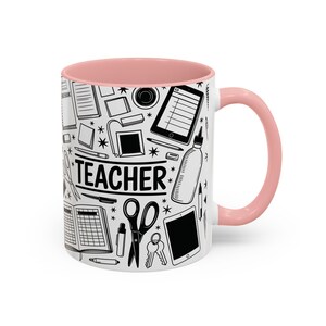 Può includere: Tazza in ceramica bianca con interno e manico rosa. La tazza presenta un design in bianco e nero con la scritta "TEACHER" e illustrazioni di materiale scolastico, tra cui forbici, tablet e quaderni.