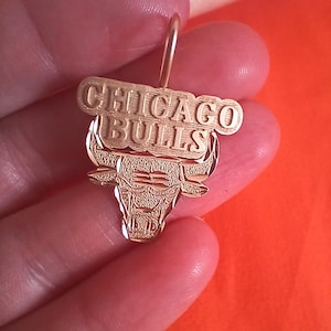May include: A gold-colored pendant featuring the Chicago Bulls logo. The pendant has the words "CHICAGO BULLS" above a detailed bull head. The design includes a loop for attaching to a chain or bracelet.