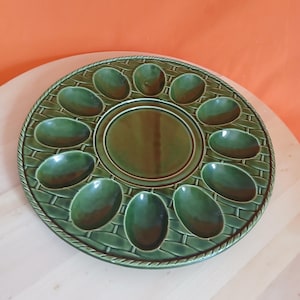 May include: A round, dark green ceramic egg serving platter featuring a central circular depression surrounded by twelve oval-shaped indentations. The surface has a textured, woven basket-weave pattern and a rope-style border around the edge.