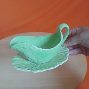 May include: A light green ceramic gravy boat with a textured leaf pattern on the exterior, featuring a curved handle and a matching oval saucer base with a scalloped edge, designed for serving sauces or dressings at the dining table.