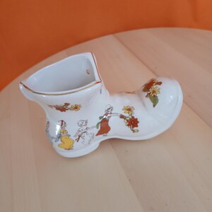 May include: A white ceramic shoe-shaped planter with a gold rim and a decorative scene of figures and flowers. The planter has a hole for hanging and is set on a light wood surface.