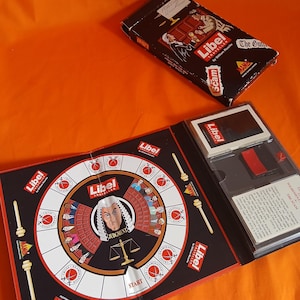 May include: A vintage "Libel Traveller" board game with a black and red design. The game includes a circular game board, cards, and a box with illustrations and text. The game board features a judge illustration and the word "Judgment".