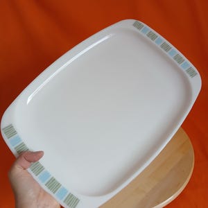 May include: A rectangular white serving tray with rounded corners. The tray has a decorative border of blue and green vertical stripes. The tray is held over a wooden surface against an orange background.