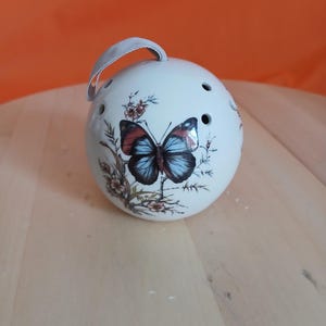 May include: A spherical white ceramic pomander featuring a detailed butterfly illustration with blue and black wings, surrounded by small floral sprigs. The object has several circular ventilation holes and a light grey fabric loop at the top.