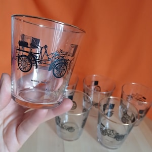 May include: Set of six clear glass tumblers. Each tumbler features a black illustration of a vintage car with the text "MORS 1908". The glasses have a classic, vintage aesthetic, suitable for serving beverages.