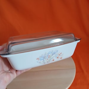 May include: A rectangular, white casserole dish with a clear glass lid. The dish features a floral design with blue and orange accents. The dish is held over a wooden surface against an orange background.