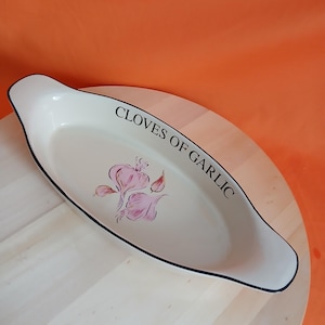 May include: Cream-coloured, oval ceramic dish with black trim and the words "CLOVES OF GARLIC". The dish features a pink garlic bulb illustration. The dish is on a wooden surface against an orange background.
