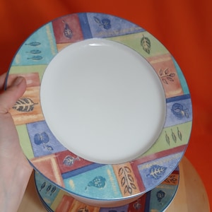 May include: A stack of white ceramic plates with a colourful border. The border features a repeating pattern of leaves, flowers, and geometric shapes in shades of blue, green, and orange. The plates are stacked on top of each other.