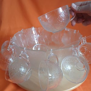 May include: A clear glass punch bowl set with a round base and several hanging cups. The bowl and cups have a leaf pattern. One hand is holding a cup above the bowl. The background is orange.