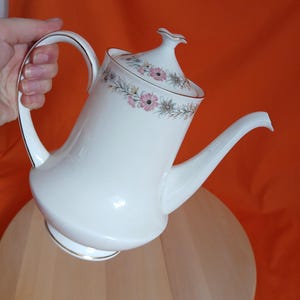 Puede incluir: Cafetera de porcelana blanca con asa y boquilla curvadas. La tapa y el borde están decorados con un estampado floral en rosa, verde y dorado. La cafetera tiene un ribete dorado y está sujeta por una mano.