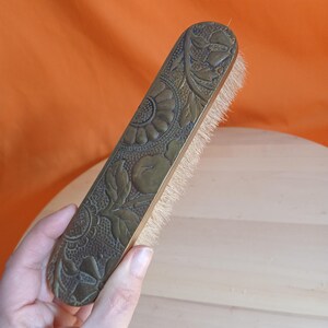May include: An antique brass brush with floral detailing. The brush has a rectangular shape with rounded ends. The bristles are light-coloured. The background is orange and light wood. The brush is held by a hand.