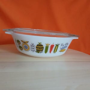 May include: White oval casserole dish with a clear glass lid. The dish is decorated with colorful illustrations of fruits and vegetables, including a pineapple, peppers, and tomatoes. The dish is on a light wood surface with an orange background.