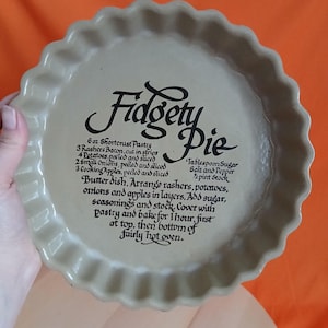 May include: A beige ceramic pie dish with a fluted edge. The dish has the words "Fidgety Pie" in a decorative font, and a recipe printed in black ink. The dish is held by a hand.