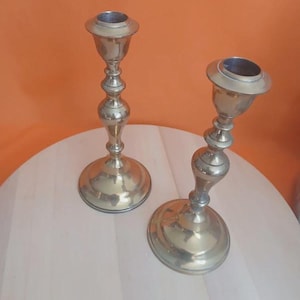May include: Two polished brass candlesticks featuring a classic turned design with a wide circular base, a slender central stem with decorative bulbous detailing, and a flared top cup designed to hold a single taper candle securely in place.