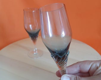 May include: Two clear champagne flutes with a dark grey and gold twisted stem design. The glasses have a classic shape with a wide bowl and a slender stem. The background is a solid orange color.