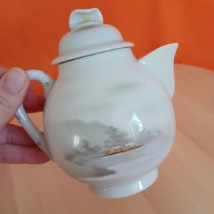 May include: Cream ceramic teapot with a landscape design. The teapot has a rounded body, a curved handle, and a spout. The lid is topped with a flower-shaped knob. The landscape design includes trees and a small structure.