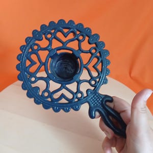 Puede incluir: Un portavelas circular de metal azul oscuro con un intrincado diseño calado de corazones y bordes festoneados. La pieza incluye una copa central circular para una vela y un mango resistente que se extiende desde el lateral para sujetarlo.
