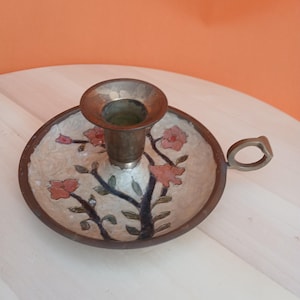 May include: A vintage brass candle holder with a floral design. The candle holder has a round base with a handle and a central cup for the candle. The base is decorated with pink flowers and green leaves.