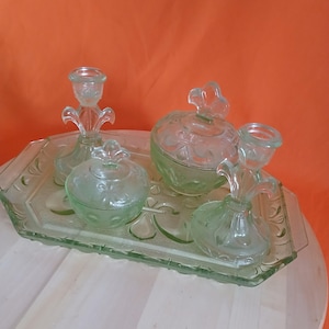 May include: A vintage green glass vanity set, including a tray, two candle holders, and two lidded containers. The set has a delicate, floral design and is displayed on a light-colored wooden surface.